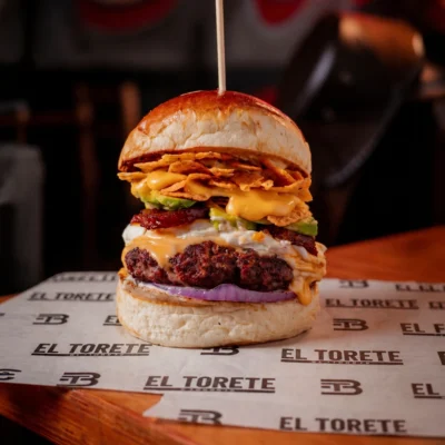 La West combina carne de res al carbón con Doritos en salsa cheddar, queso cheddar, cremoso de queso Philadelphia, aguacate, maduros caramelizados, cebolla morada y salsa tártara en pan de mantequilla, acompañada de papas crocantes.